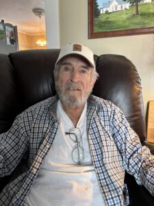 Man in his seventies sitting in a chair with a cap on.