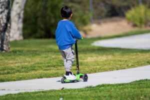 A young boy riding a green scooter on a sunny day in the park.