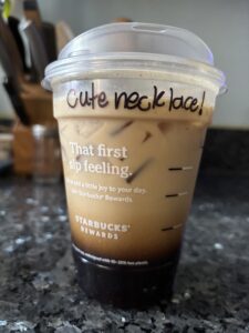 A starbucks clear coffee cup sits on a counter with the words "cute necklace" written on the side.