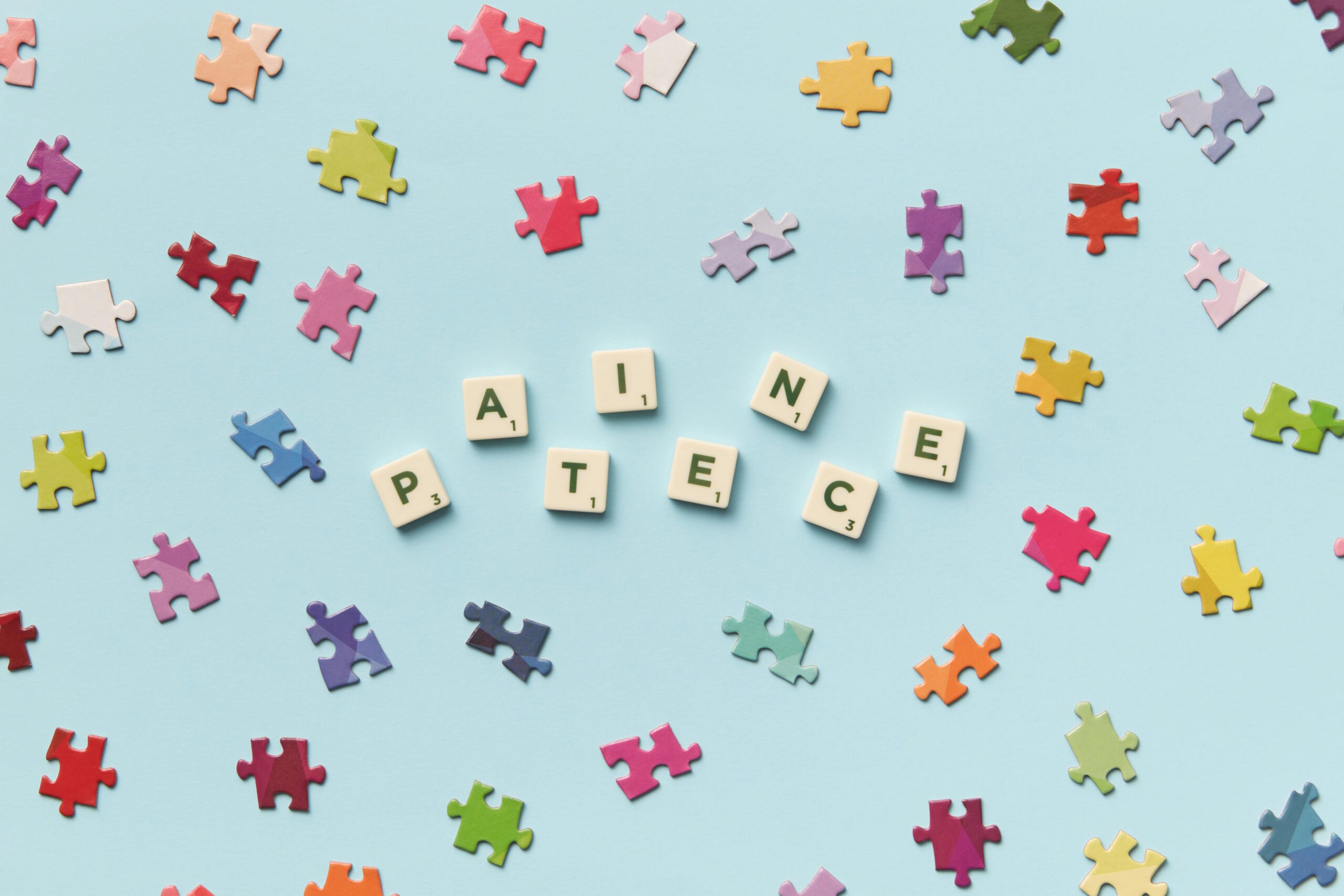 Puzzle pieces scattered on a blue surface with the word patience spaced out in scrabble letters in the middle.